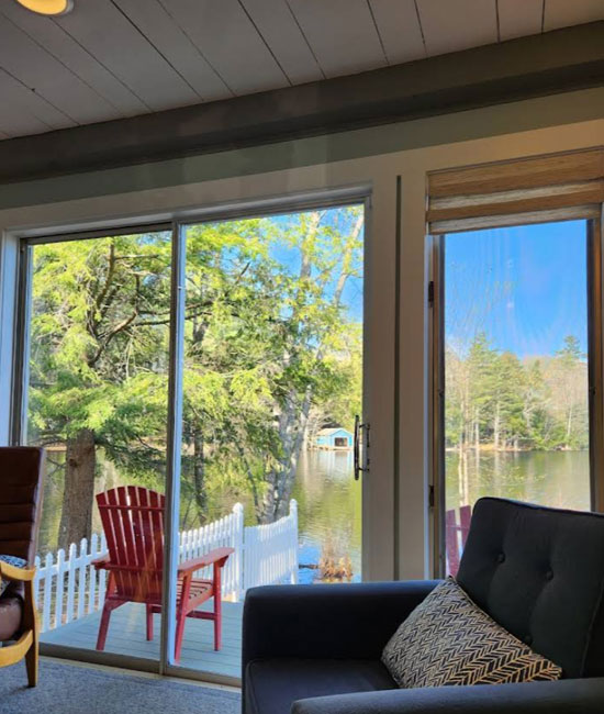 The image shows a spacious living room with a large patio door offering a view of a serene lake and trees, furnished with a couch and chair, and a cozy cabin-like atmosphere.