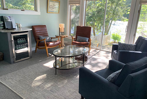 The image shows an interior view of a spacious living room with large windows offering a scenic outdoor view, featuring furniture such as sofas, chairs, and a coffee table, along with decorative elements like rugs and artwork, and a modern kitchenette in the background with a countertop and stools.