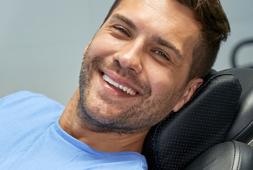 The image shows a man sitting on a dental chair with his feet up, smiling broadly at the camera, wearing a blue shirt.