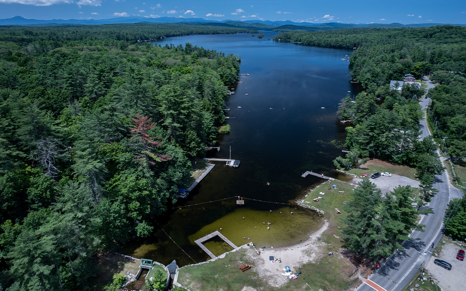 The image shows a scenic outdoor area with a large body of water, likely a lake, surrounded by lush greenery and trees, featuring a dock on the left side with boats and people enjoying the water, and a sandy beach area on the right, with a clear blue sky above.
